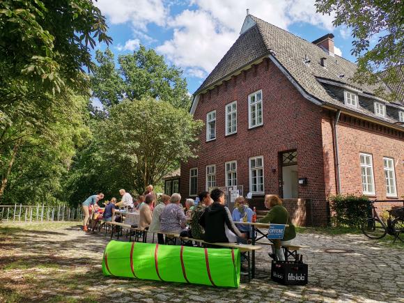 Nachbar:innen sitzen zusammen an einer langen Tafel vor einem alten Kasernengebäude. Die Nachbarschaft ist gemischt: Jung und alt, weiß und nicht-weiß. Vor der langen Tafel steht ein Spieltunnel für kleine Kinder zum Durchkriechen